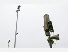 Ein hoher Lautsprechermast mit mehreren Lautsprechern und Verstärkereinheiten im Vordergrund, daneben ein separater Mast mit Flutlichtstrahlern vor bewölktem Himmel.
