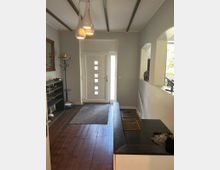 Eingangsbereich eines Hauses mit einer weißen Tür, rechteckigen Glasausschnitten und einem schmalen seitlichen Fenster. Der Raum hat einen Holzfußboden, eine Garderobe an der Wand, eine Sitzbank mit Körben und eine moderne Pendelleuchte an der Decke.