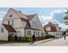 Straßenszene in einer Wohngegend mit zwei weißen Häusern im traditionellen Stil, jeweils mit roten Ziegeldächern und markanten Giebeln. Im Vordergrund führt ein gepflasterter Gehweg entlang eines niedrigen Holzzauns, eine Person geht zu Fuß, während eine andere mit dem Fahrrad fährt.