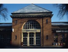 Ziegelfassade eines historischen Bahnhofsgebäudes mit dem Schriftzug „Sonnenallee“ über einem Eingang mit weiß gerahmten Türen und einer großen Bogenfensterreihe. Das Dach ist leicht mit Schnee bedeckt, und vor dem Gebäude steht eine Uhr sowie ein Mülleimer.