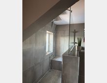 Modernes Badezimmer mit bodengleicher Dusche, grauen Fliesen und Glasabtrennung. Ein Fenster bringt Tageslicht herein, und eine Zimmerpflanze steht auf einer breiten Ablage.