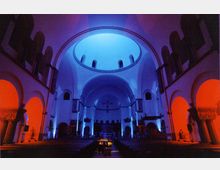 Innenansicht einer Kirche mit hohen Rundbögen und einer Kuppel, die in blauem und rotem Licht beleuchtet ist. Im Vordergrund befinden sich Kirchenbänke, während im Hintergrund ein Altar und ein großes Kreuz zu sehen sind.