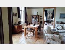 Wohnzimmer mit antikem Flair, ausgestattet mit einem Holzesstisch, Stühlen, einem Kerzenständer und einem großen Schrank. Der Raum hat helle Wände, Holzböden, Polstermöbel mit floralen Mustern und einen Durchgang zu einem weiteren Raum mit Bücherregalen.