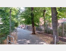 Gepflasterter Gehweg mit Bäumen und einem Metallzaun auf der rechten Seite, dahinter ein Gebäude aus rotem Backstein. Auf der linken Seite stehen ein Laternenpfahl und Fahrradständer, im Hintergrund sind Personen und weitere Gebäude erkennbar.
