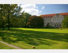 Weitläufige Parklandschaft mit grünem Rasen, vereinzelten großen Bäumen und einem historischen Gebäude mit rotem Dach und klassizistischer Fassade im Hintergrund; Personen entspannen auf der Wiese bei Sonnenschein.