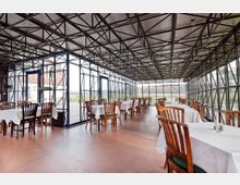 Ein Innenraum eines verglasten Restaurants mit Metallstruktur-Dachkonstruktion. Der Raum ist mit Holztischen und Stühlen bestückt, die weiß eingedeckt sind, und bietet durch die Glaswände einen Blick auf die ländliche Umgebung.