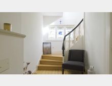 Heller Flur mit Holztreppe, dekorativem Sprossengeländer und einem großen, verzierten Fenster mit bunten Glasscheiben. Vor der Treppe steht ein schwarzer Sessel neben einem kleinen weißen Ziergefäß.