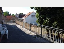 Große Steinterrasse mit einem Metallgeländer, das teilweise rostig ist, und mehreren weißen Plastikstühlen entlang einer Wand. Im Hintergrund sind Wohnhäuser mit roten Ziegeldächern und ein großer Baum zu sehen.