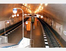 Unterirdischer U-Bahnhof mit gebogener Decke aus weißen Kacheln, beleuchtet von kugelförmigen Lampen. Der Bahnhof hat zwei Gleise mit einem Mittelbahnsteig und Treppenaufgängen, sowie Werbeplakate an den Wänden.
