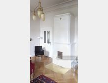 Wohnzimmer mit einem weißen Kachelofen, einer schräg gelegten Metallplatte davor und einem hölzernen Parkettboden. Im Raum stehen ein schwarzer Sessel, eine Stehlampe und ein roter Teppich; eine weiße Tür mit Glaseinsätzen ist an der Wand sichtbar.