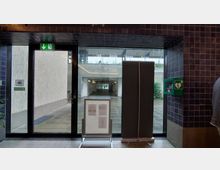 Eine Glastür in einem Gebäude mit Blick auf einen überdachten Durchgang und eine Außenfläche. Links an der Tür ein Notausgangsschild, rechts ein Defibrillatorkasten, davor ein Infoständer und eine Trennwand.