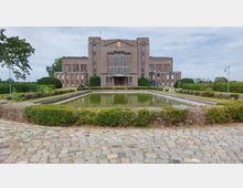 Backsteingebäude im Stil der frühen Moderne mit symmetrischer Fassade und großen Fenstern, davor ein rechteckiges Wasserbecken, umgeben von gepflegten Hecken und einem gepflasterten Platz. Im Hintergrund ländliche Umgebung mit vereinzelten Bäumen.