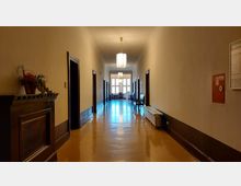 Ein langer, heller Korridor in einem Gebäude mit glänzendem Holzboden und beidseitigen Holztüren. Am Ende des Flurs befindet sich ein großes Fenster, darüber hängen moderne Hängelampen, und an der rechten Wand sind Sitzbänke sowie ein Feuerlöschplan angebracht.