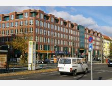 Rote Backsteinfassade eines mehrstöckigen Büro- und Geschäftsgebäudes mit großen Fenstern und Läden im Erdgeschoss, darunter eine Bäckerei, ein Supermarkt und ein Videogeschäft. Im Vordergrund verlaufen eine Straße mit parkendem und fahrendem Verkehr sowie ein Gehweg mit einer Bushaltestelle.