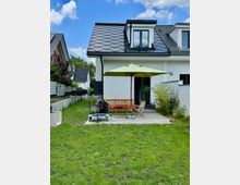 Ein modernes Einfamilienhaus mit dunklem Satteldach und Dachgauben, im Vordergrund ein Garten mit Terrasse. Auf der Terrasse stehen Gartenmöbel, ein Sonnenschirm und ein kleines Planschbecken auf Rasen.