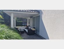 Überdachte Terrasse eines modernen Hauses mit einer weißen Ecksofa-Garnitur und Glastisch, umgeben von hohen Gräsern und einem gepflegten Garten mit Hecken. Im Hintergrund ist ein Trampolin sichtbar.