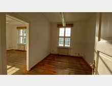 Ein leerer Innenraum mit Holzboden, weißen Wänden und zwei Fenstern mit Sprossen, die Tageslicht hineinlassen. Im Vordergrund ist eine geöffnete Tür sichtbar, und unter den Fenstern befindet sich ein Heizkörper.