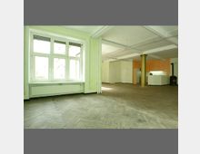 Großer, heller Innenraum mit Parkettboden in Fischgrätenmuster, weiß gestrichenen Wänden und freiliegenden Deckenbalken. Der Raum verfügt über ein großes Fenster, eine offene Küche mit weißen Schränken und einen freistehenden Ofen in einer Ecke.