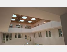 Innenansicht eines modernen, mehrstöckigen Atriums mit umlaufenden Galerien, weißen Wänden und vielen rechteckigen Fenstern. In der hohen Decke sind mehrere große, runde Oberlichter eingelassen, ergänzt durch eingelassene Spotbeleuchtung entlang der Galerie.