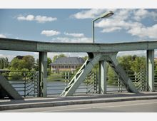 Grüne Stahlbrücke mit Nietenkonstruktion und verzierten Geländern, im Hintergrund ein historisches Gebäude mit grauem Dach, umgeben von Bäumen und einem Fluss. Der Himmel ist leicht bewölkt.
