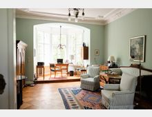 Wohnzimmer mit klassischem Interieur, grünen Wänden und ornamentierter Decke. Im Vordergrund stehen zwei gestreifte Sessel und ein bunter Teppich, im Hintergrund ein Erker mit großen Fenstern, Holzmöbeln und einem Schreibtisch.