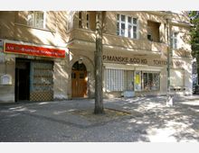 Fassade eines Wohn- und Geschäftsgebäudes in einer Stadt mit einem Ladeneingang "Am S-Bahnhof Sonnenallee" und Schaufenstern des Geschäfts "P. Manske & Co KG - Tortenversand". Vor dem Gebäude stehen ein Baum und eine Straßenlaterne, gepflasterter Gehweg darunter.