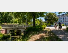 Uferweg an einem kleinen Bach mit einer niedrigen Betonbrücke und Metallgeländer, umgeben von dichtem Grün und großen Bäumen. Rechts verläuft ein Schotterweg neben einer Straße, im Hintergrund steht ein mehrstöckiges, helles Gebäude mit vielen Fenstern.