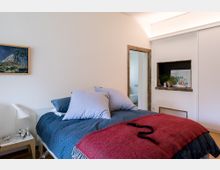 Schlafzimmer mit einem Doppelbett, das mit blauer Bettwäsche, weißen Kissen und einer roten Decke ausgestattet ist. Im Hintergrund befinden sich eine Tür zu einem angrenzenden Raum, ein kleines Wandregal mit Pflanzen sowie ein Gemälde an der weißen Wand.