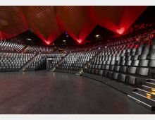 Moderner Veranstaltungssaal mit ansteigenden Sitzreihen aus schwarzen Stühlen, beleuchteten Treppenstufen und einem auffälligen geometrischen Deckendesign in Rot- und Orangetönen. Der Raum wirkt groß und ist für Zuschauertribünen ausgelegt.