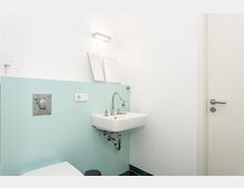 Kleines, helles Badezimmer mit wandmontiertem Waschbecken, verchromter Armatur und Mintgrün-Fliesen als Wandverkleidung. Über dem Becken hängt eine rechteckige Wandleuchte, daneben ist eine weiße Tür zu sehen.