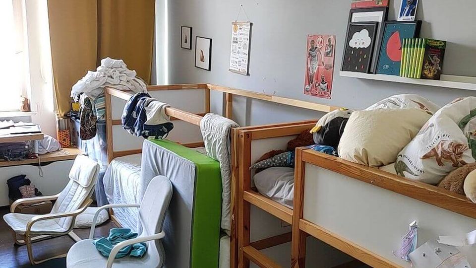 A children’s bedroom with wooden bunk beds against a gray wall, decorated with books, posters, and small wall art. The room features a window with mustard yellow curtains, a white cushioned chair, and scattered toys and books on a blue carpeted floor.