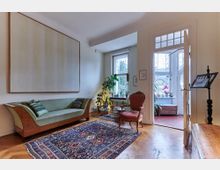 Wohnzimmer mit Holzboden, einem großen, gemusterten Teppich und einer hellgrünen Couch. Im Raum stehen zudem ein antiker roter Sessel und Pflanzen vor zwei großen Fenstern, die Tageslicht in den Raum lassen.