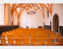 Innenansicht eines historischen Veranstaltungsraums mit weißen Wänden und einem Deckengewölbe aus roten Ziegelbögen. Der Raum ist mit mehreren Reihen gelber Holzstühle ausgestattet, großen Bogenfenstern an der linken Seite und einer Tür mit Steinrahmen auf der rechten Seite.