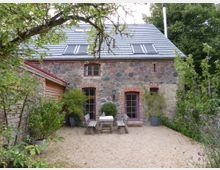 Steinhaus mit rustikaler Fassade aus gemischtem Naturstein und roten Backsteindetails, grauem Dach mit Dachfenstern, umgeben von einem Garten mit Bäumen und blühenden Pflanzen. Im Vordergrund ein kleiner Kiesplatz mit Holztisch, Sitzbänken und Pflanzkübeln.