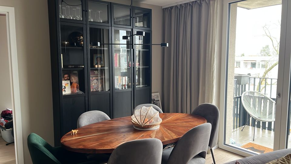 Essbereich in einer modernen Wohnung mit ovalem Holztisch und mehreren Polsterstühlen vor einem hohen schwarzen Vitrinenschrank. Rechts führen bodentiefe Glastüren mit grauen Vorhängen auf einen Balkon mit Metallgeländer und einem weißen Outdoor-Stuhl.