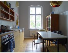 Helle Küche mit Holzboden, einem weißen Esstisch und unterschiedlichen Stühlen. Links befinden sich eine Arbeitsplatte mit Herd und Spüle sowie hängende Holzregale, rechts ein Holzschrank, vor einem großen Fenster mit Blick ins Grüne.