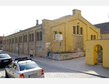 Gelbes Backsteingebäude mit Reihen von hohen Fenstern und einem großen Rolltor, beschriftet als „Reithalle B“, mit Graffiti auf den Wänden; im Vordergrund parkende Autos und eine kleine Mauer mit Sitzenden. Ein gelber Torbogen mit Gitter befindet sich rechts.