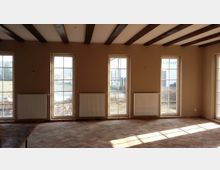 Heller Innenraum mit großen bodentiefen Fenstern, beige Wänden und einem Boden aus roten Ziegelsteinen in Fischgrätmuster; an den Wänden sind weiße Heizkörper angebracht, und die Decke ist mit dunklen Holzbalken versehen.