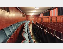 Innenraum eines Theaters oder Auditoriums mit gepolsterten Sitzreihen in Blau und Holzvertäfelung an den Wänden. Die Decke ist schlicht mit rechteckigen, deckenbündigen Leuchten, und mehrere geschlossene Türen befinden sich an der Rückwand.