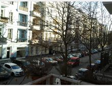 Straßenszene in einem städtischen Wohngebiet mit mehrstöckigen Altbauhäusern in Pastellfarben, Balkonen und großen Fenstern. Im Vordergrund stehen kahlen Bäume, darunter geparkte Autos entlang der Straße.