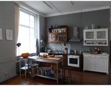 Wohnküche mit industriellem Charme, grauen Wänden und großen Fenstern, die viel Tageslicht hereinlassen. Die Einrichtung umfasst eine Küchenzeile mit Fliesenrückwand im bunten Muster, einen weißen Schrank, eine Dunstabzugshaube, einen Herd sowie einen freistehenden Holztisch mit Kochutensilien.