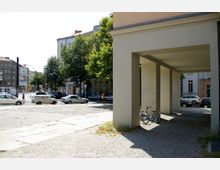 Straßenszene mit modernen Wohngebäuden im Hintergrund, geparkten Autos, Bäumen entlang der Straße und einer überdachten Passage auf der rechten Seite mit einem Kinderfahrrad abgestellt auf gepflastertem Boden.