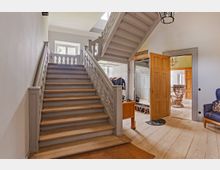 Treppenhaus in einem hellen Innenraum mit einer hölzernen Treppe, die auf beiden Seiten Geländer aus geschnitztem Holz hat. Im Hintergrund befinden sich offene Türen, durch die angrenzende Zimmer mit Holzmöbeln und Dekorationselementen sichtbar sind.