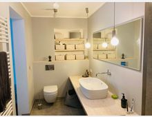 Modernes Badezimmer mit weißen Wänden und grauem Bodenfliesen. Zu sehen sind ein wandmontiertes WC, ein Holzwaschtisch mit ovalem Waschbecken, ein großer Wandspiegel, hängende Lampen sowie offene Regale mit geflochtenen Körben.