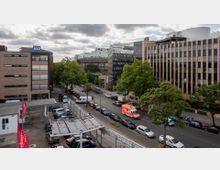 Straßenszene in einer Stadt mit mehreren modernen Bürogebäuden, teils mit Glasfassaden. Entlang der Straße befinden sich geparkte Autos, ein Krankenwagen und Bäume, eine Tankstelle ist im Vordergrund zu sehen.