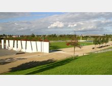 Ein moderner Park mit breiten Gehwegen, einer Wiese mit Fußballtoren und einem überdachten Bereich im Hintergrund. Weiße, geneigte Betonwände auf einer asphaltierten Fläche dominieren den Vordergrund, umgeben von Bäumen und Grünflächen.