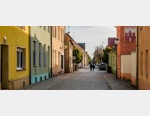 Pflasterstraße in einem historischen Wohnviertel mit farbenfrohen, einfachen Gebäuden in Gelb, Grün und Orange. Zwei Personen spazieren die Straße entlang, die von kleinen Bäumen und vereinzelten Laternen gesäumt ist.