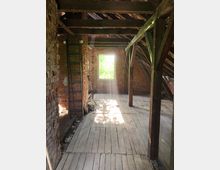 Dachboden eines alten Gebäudes mit freiliegenden Ziegelwänden, Holzbalken und Holzboden. Ein Fenster, durch das Sonnenlicht einfällt, beleuchtet den Raum; eine Leiter lehnt an der linken Wand.