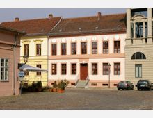 Eine historische Platzszene mit mehrere Gebäuden in Pastellfarben; ein gelbes, ein rosa Gebäude mit roten Fensterrahmen und dekorativen Stuckdetails, sowie ein moderneres Gebäude mit Sandsteinfassade. Vor den Gebäuden stehen Autos, Töpfe mit Pflanzen und eine Straßenlaterne auf einem mit Kopfsteinpflaster bedeckten Platz.
