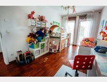Kinderzimmer mit hellem Holzboden, einem halbhohen Bett mit dekorativem Stoffvorhang und Spielmotiven, reichlich Regalen voller Spielzeug und Büchern sowie einem Schreibtisch mit rotem Drehstuhl. Große Fenster mit weißen Vorhängen und Jalousien lassen viel Tageslicht herein.